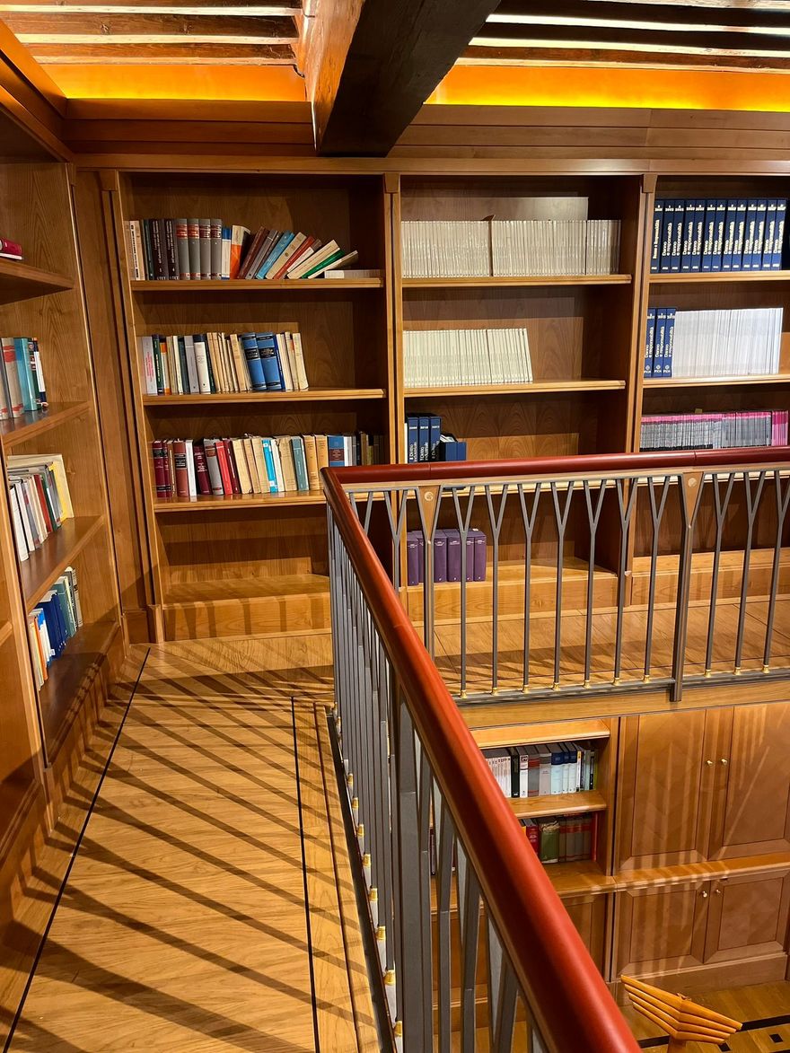 Biblioteca in legno con libri su scaffali, scale e ringhiera. Illuminazione calda e atmosfera accogliente.