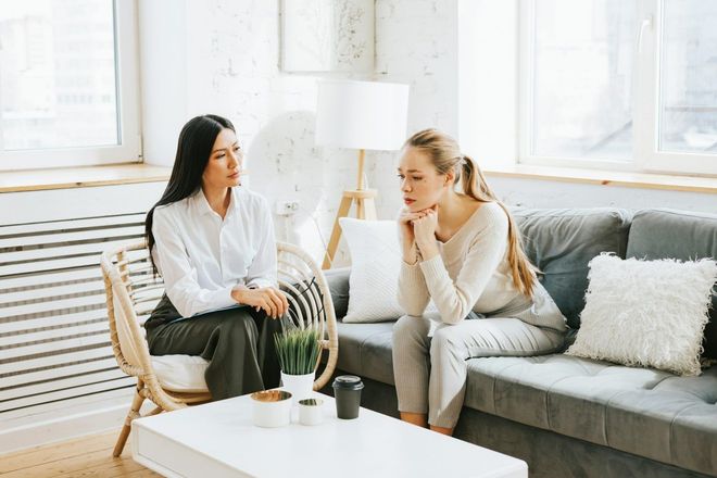 Two women sit, one in a chair, the other on a couch. They appear to be in a serious discussion, in a well-lit living room.