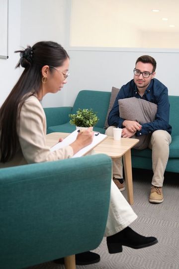 Therapist with notepad listens to a person holding a pillow on a teal couch; coffee table in between.