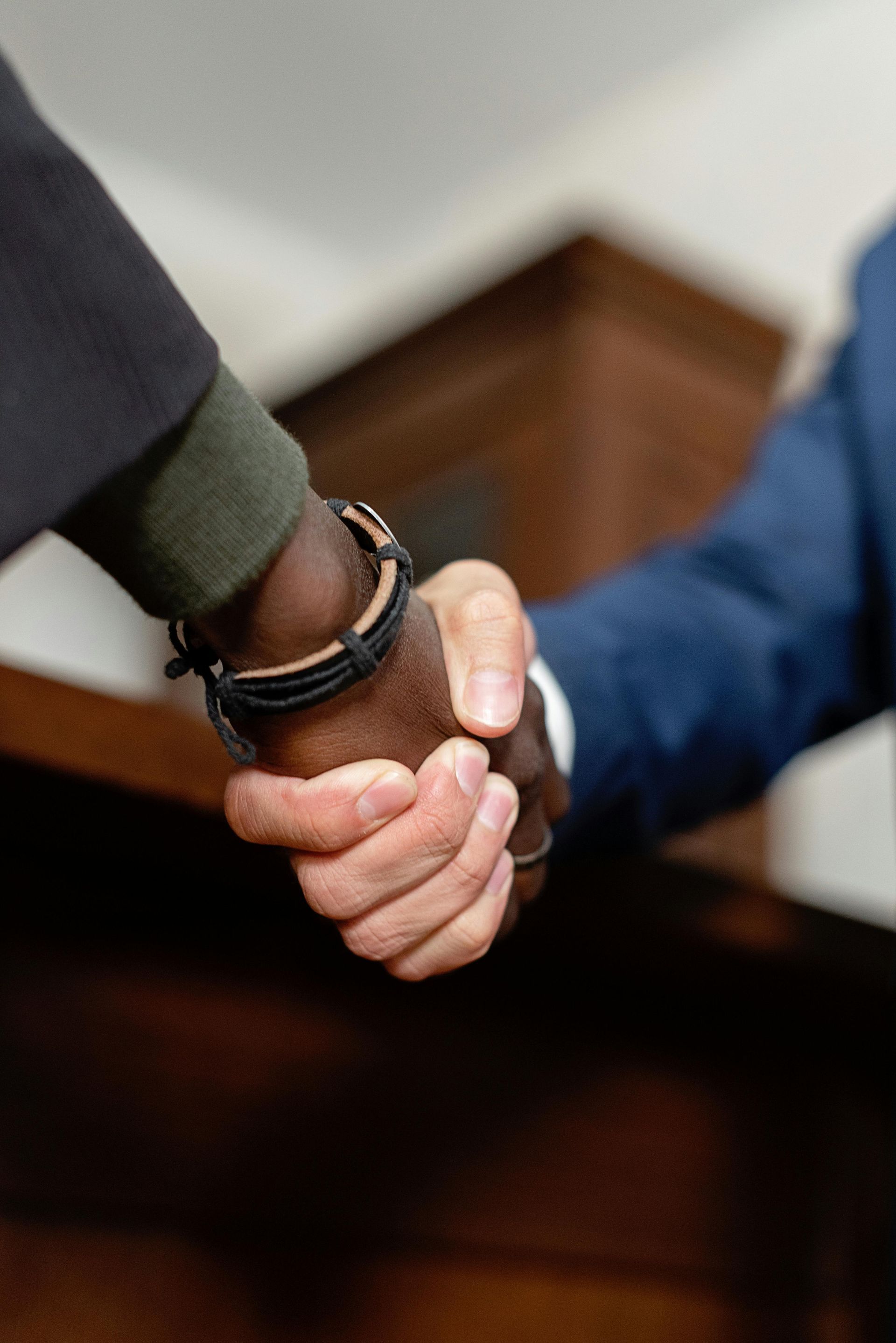 Two people shaking hands over a dark wooden table, one wearing a suit and the other a watch.