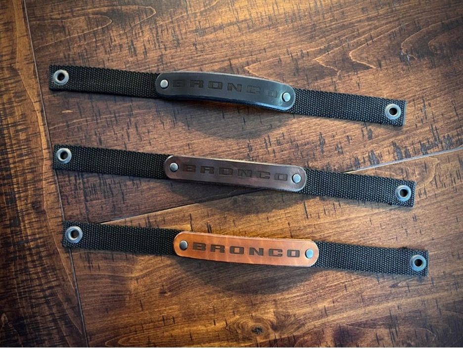 Three leather Bronco wristbands on a wood table, each with the word "BRONCO" in a different color.
