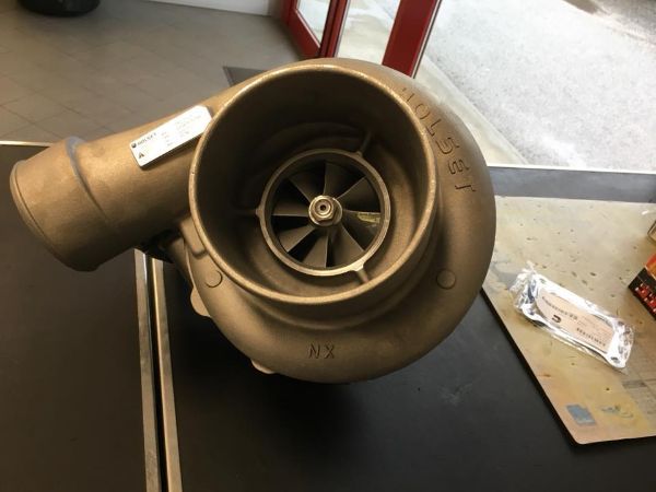 Un turbocompressore metallico con una pala della ventola a vista, appoggiato su un piano di lavoro accanto a un piccolo componente confezionato.