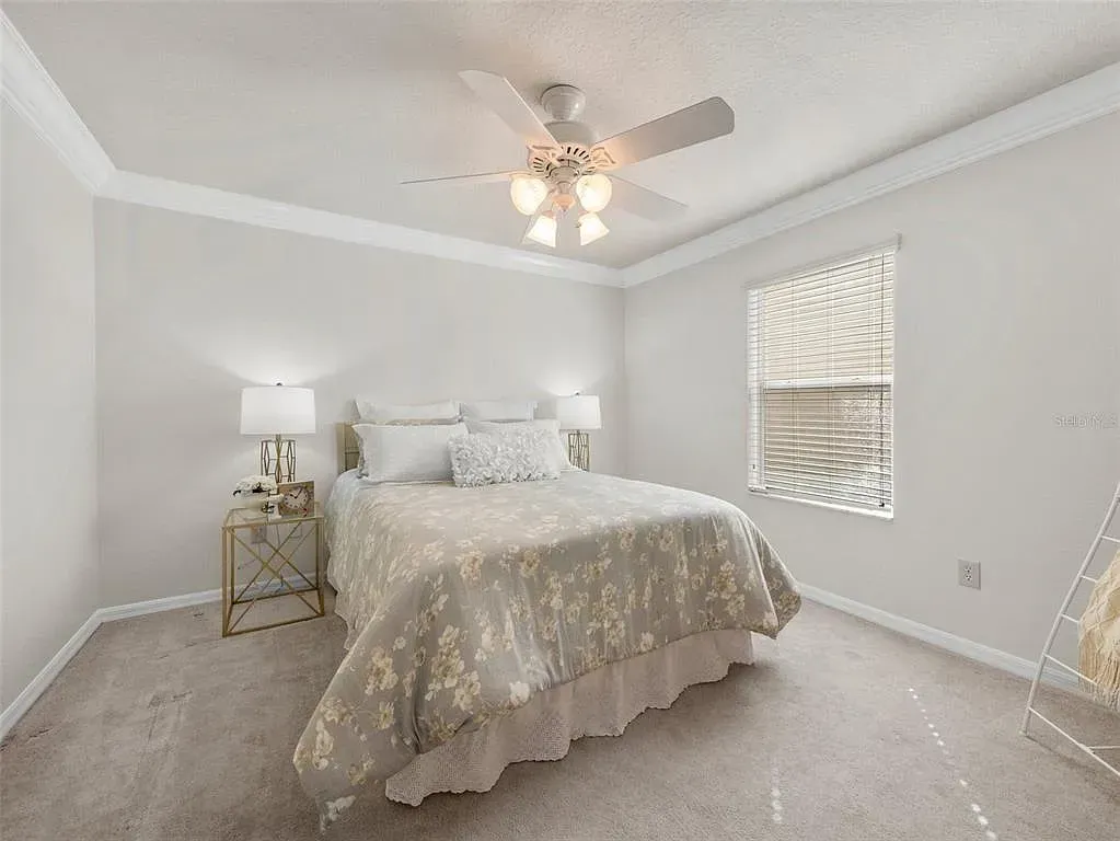 Bright bedroom with a bed, ceiling fan, bedside table, and window blinds on light gray walls.