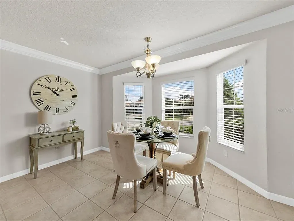 Bright dining room with round table, cream chairs, large wall clock, and bay windows with a chandelier.