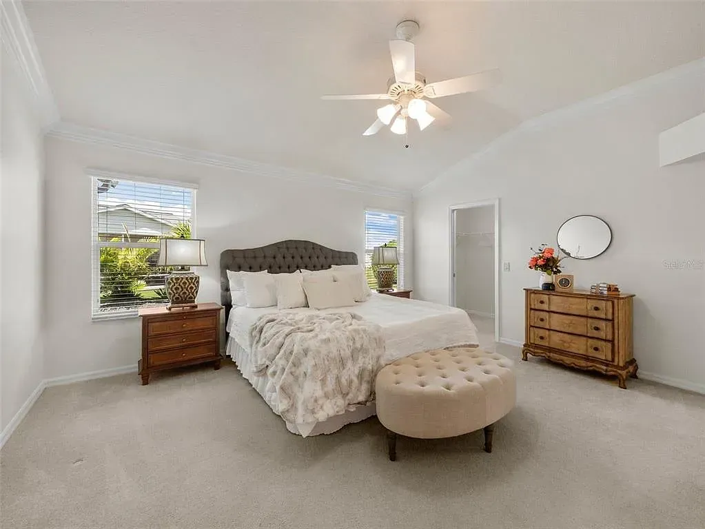 Bright white bedroom with a bed, nightstands, tufted bench, and ceiling fan near sunlit windows
