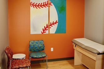 Waiting room with orange walls, two chairs, abstract artwork, and an exam table