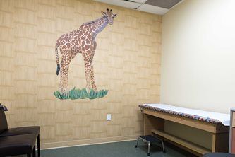 Exam room with giraffe mural, exam table, and chair against beige walls