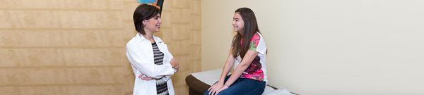 Two women talking in a bright room, one seated on a treatment table in a clinic.