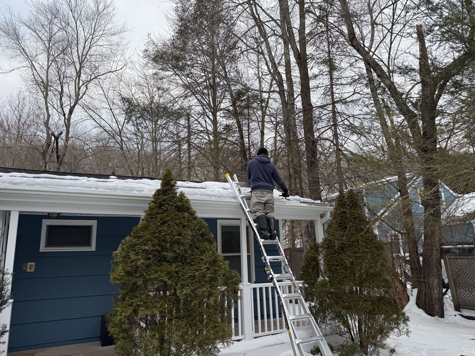 Person on a ladder clearing snow from a house roof in a winter yard
