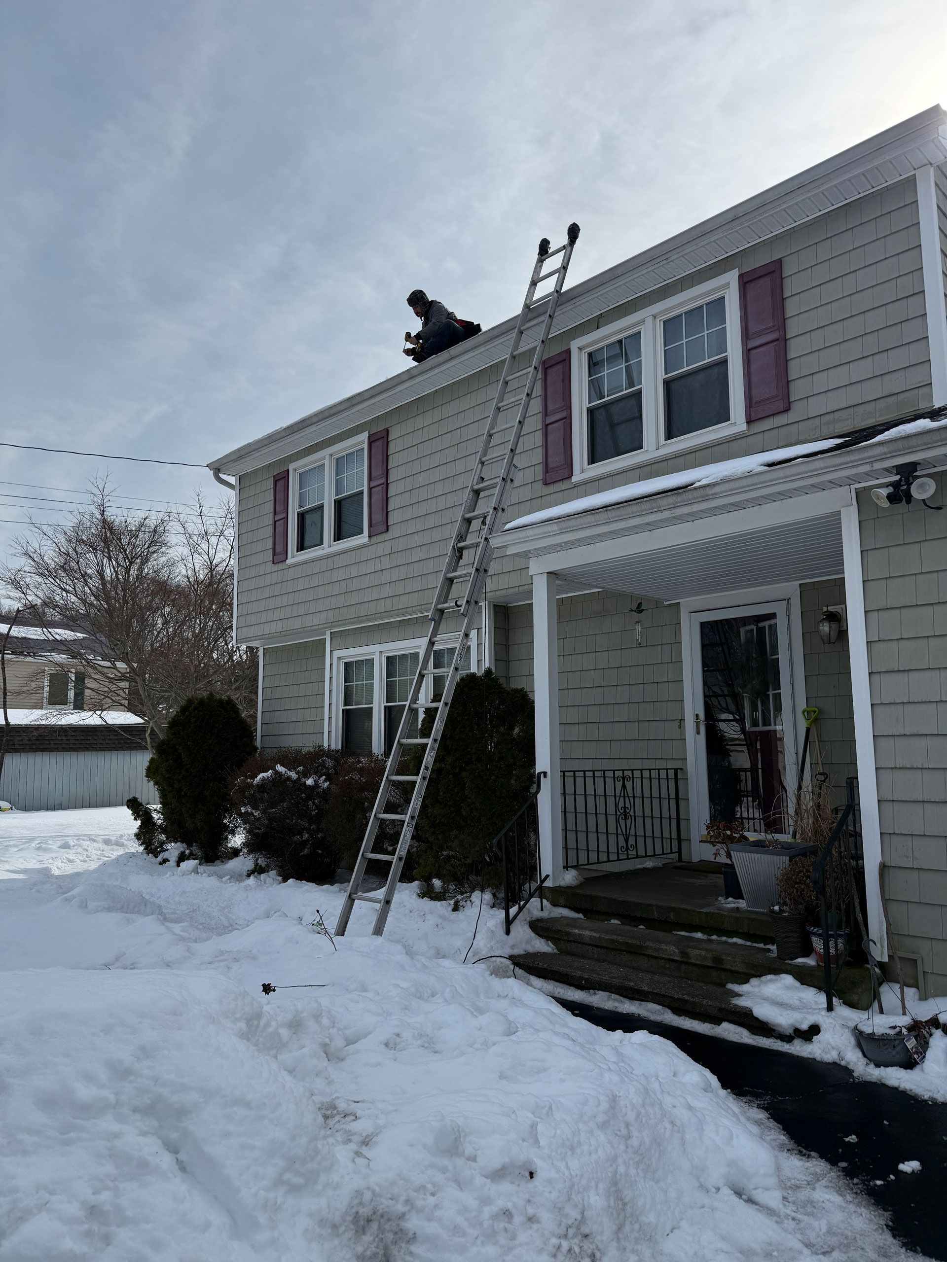 Worker on extension ladder inspecting roof edge of a gray house under a blue sky