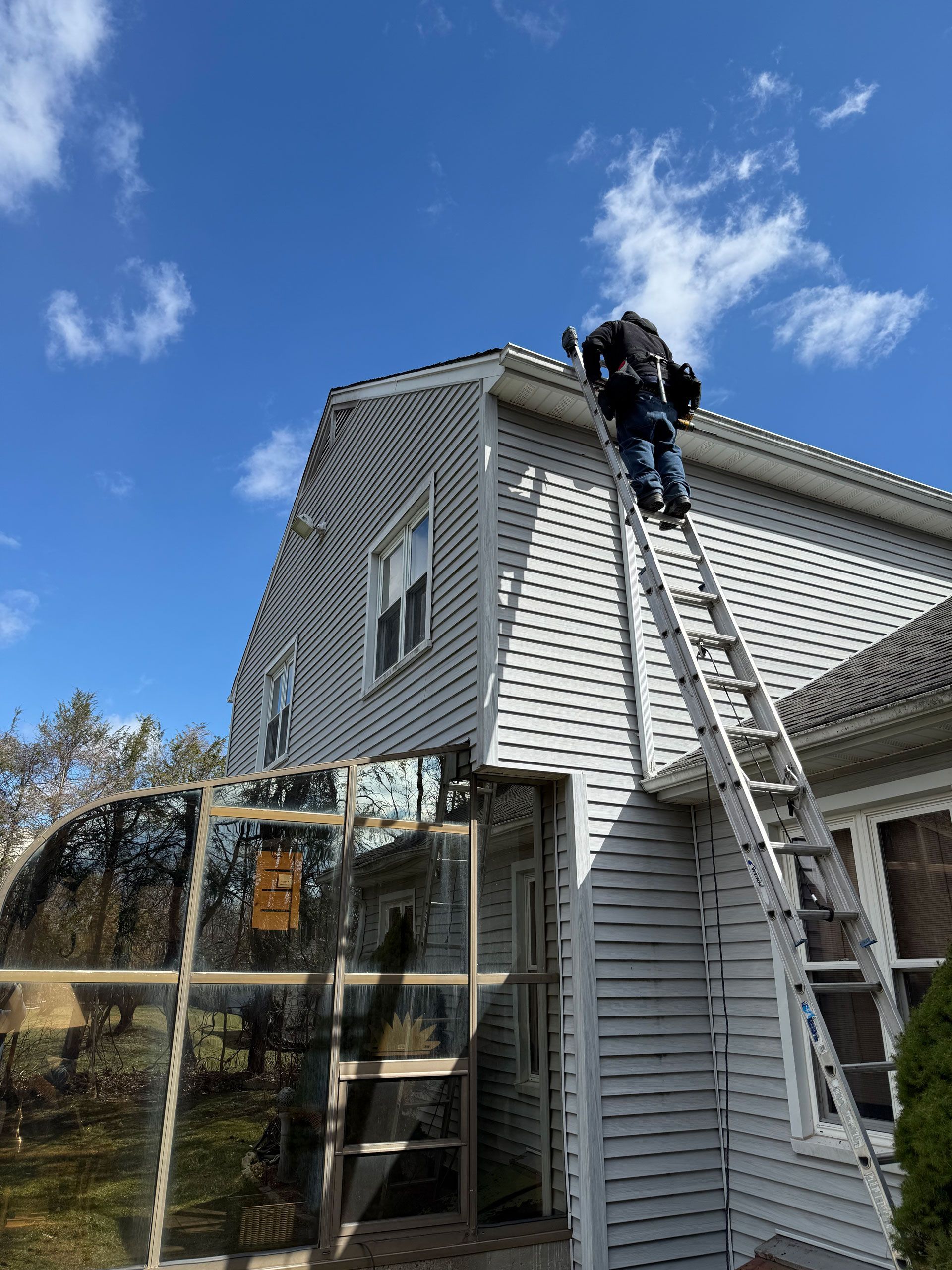 Worker on a ladder cleaning gutters on a white house under a blue sky.