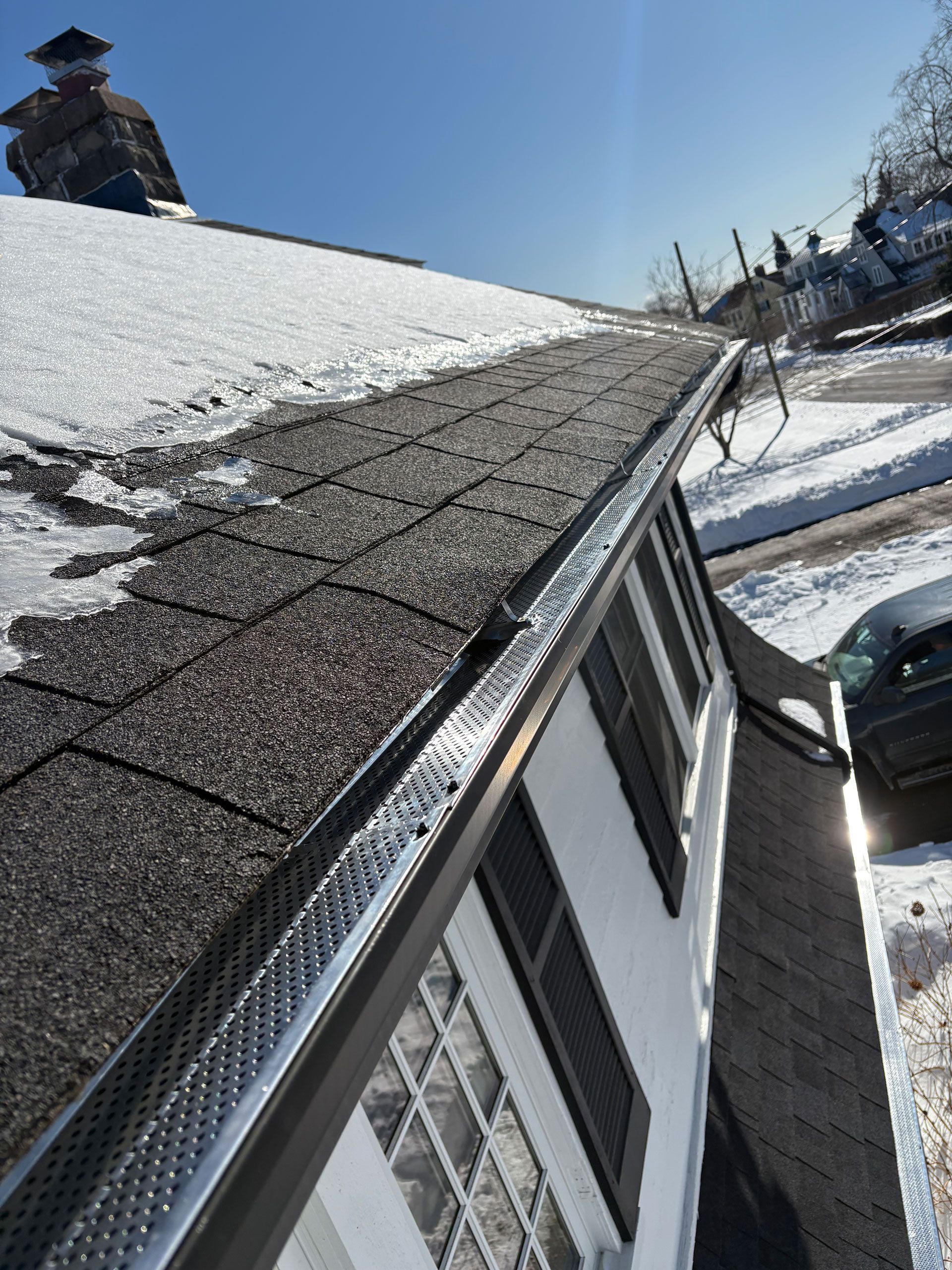 Snow-covered house roof with shingles and icicles beside a snowy yard on a sunny winter day