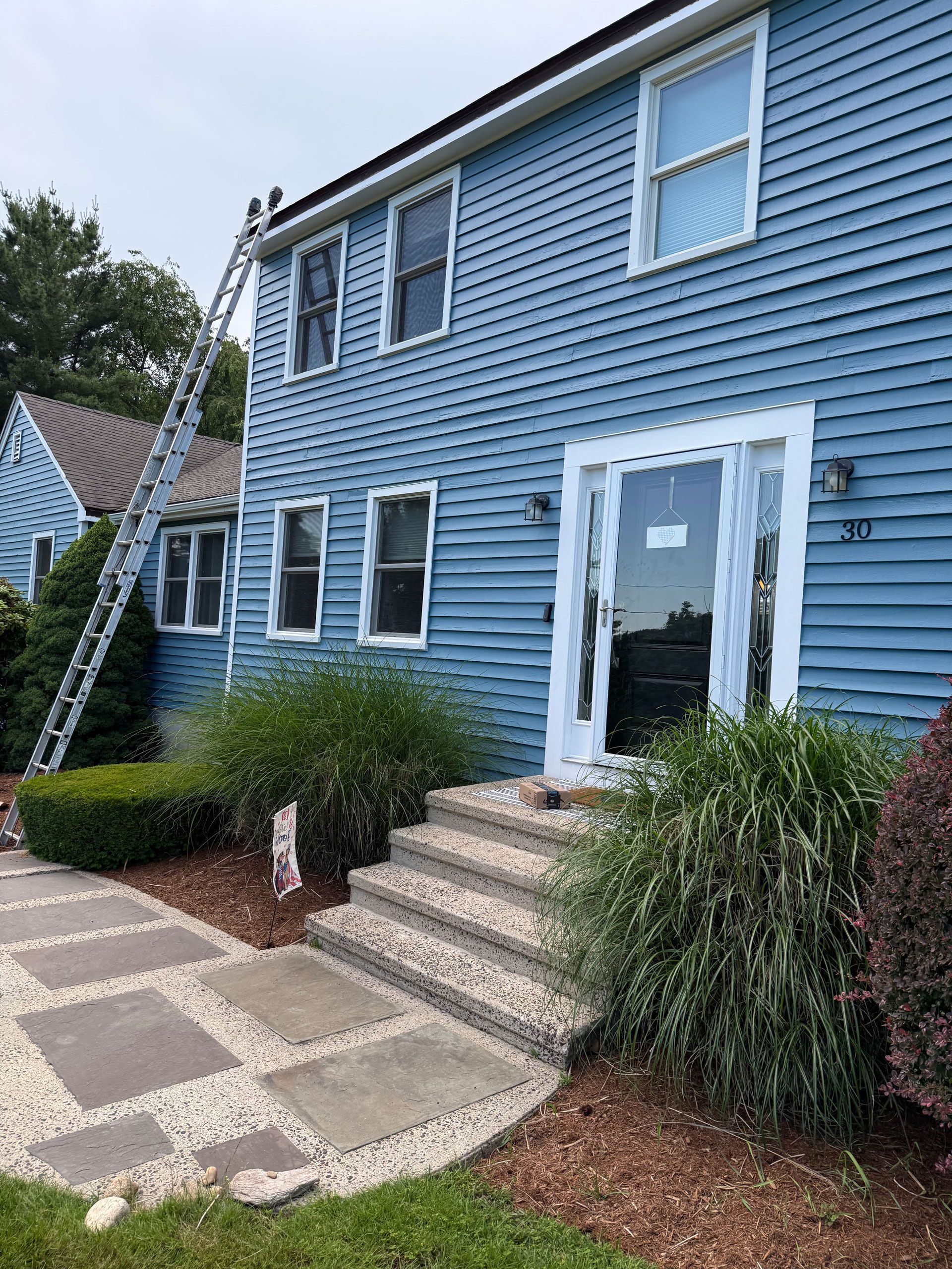Blue shingle-sided house with white trim, front steps, and a glass door beside a ladder and shrubs.