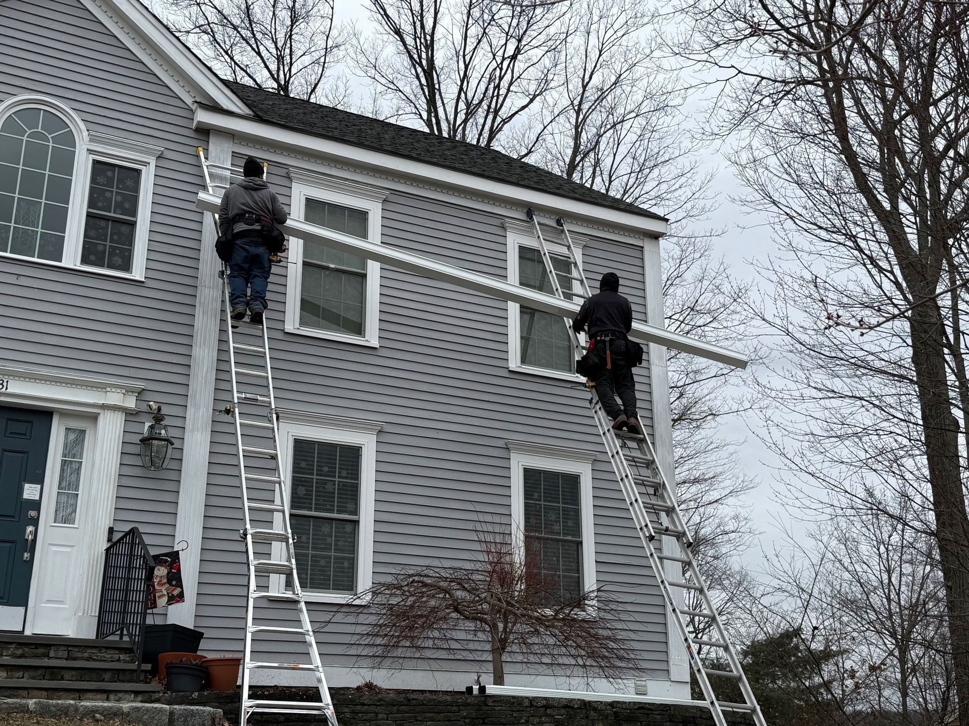 Two workers on ladders painting the exterior of a gray house in winter.