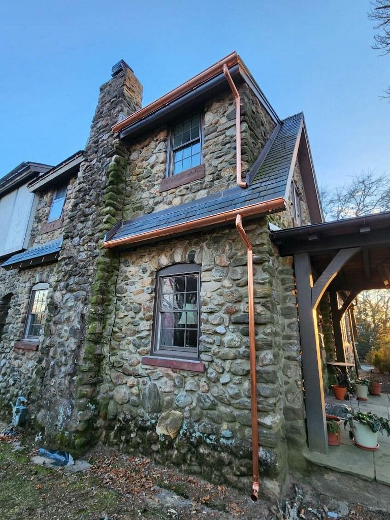 Stone cottage exterior with red-trimmed roof and window, beside a covered porch and chimney.