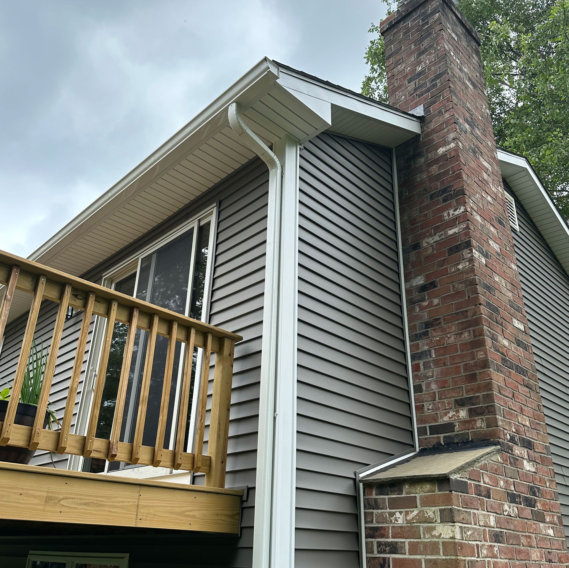 Exterior of a house with gray siding, wooden deck railing, and a large brick chimney under cloudy sky