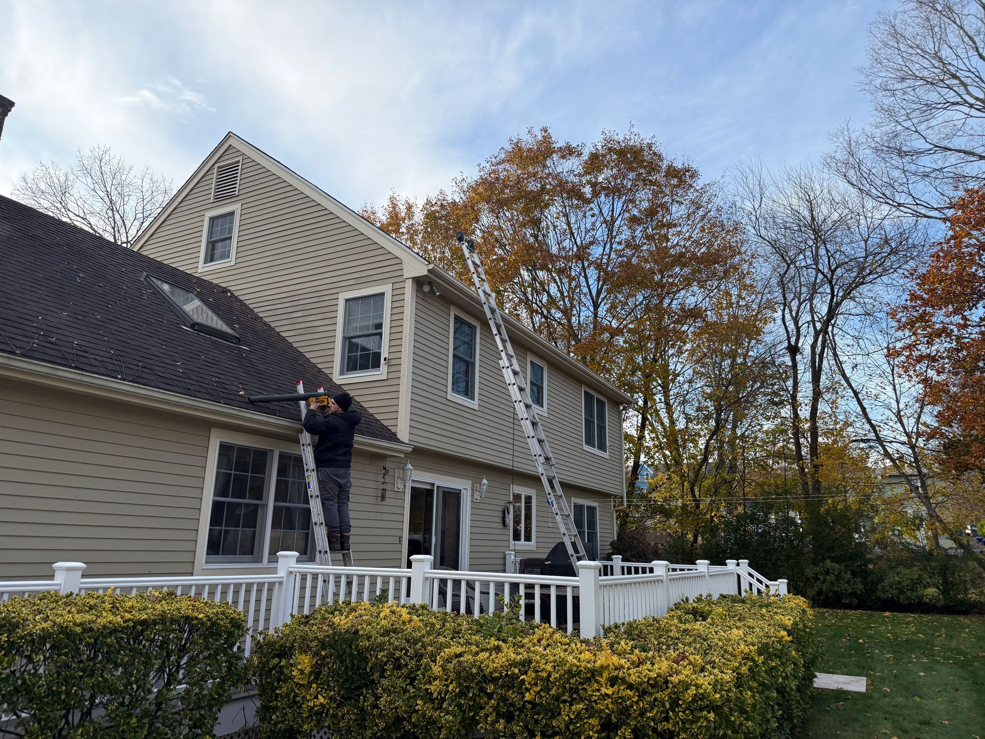 Person on a ladder painting a beige house with white trim and a porch, surrounded by autumn trees