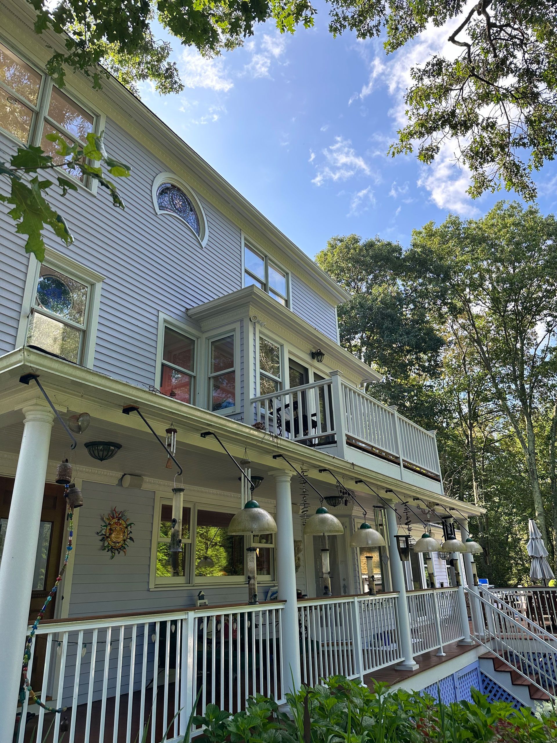 Two-story white house with porch and balcony, surrounded by trees under a blue sky