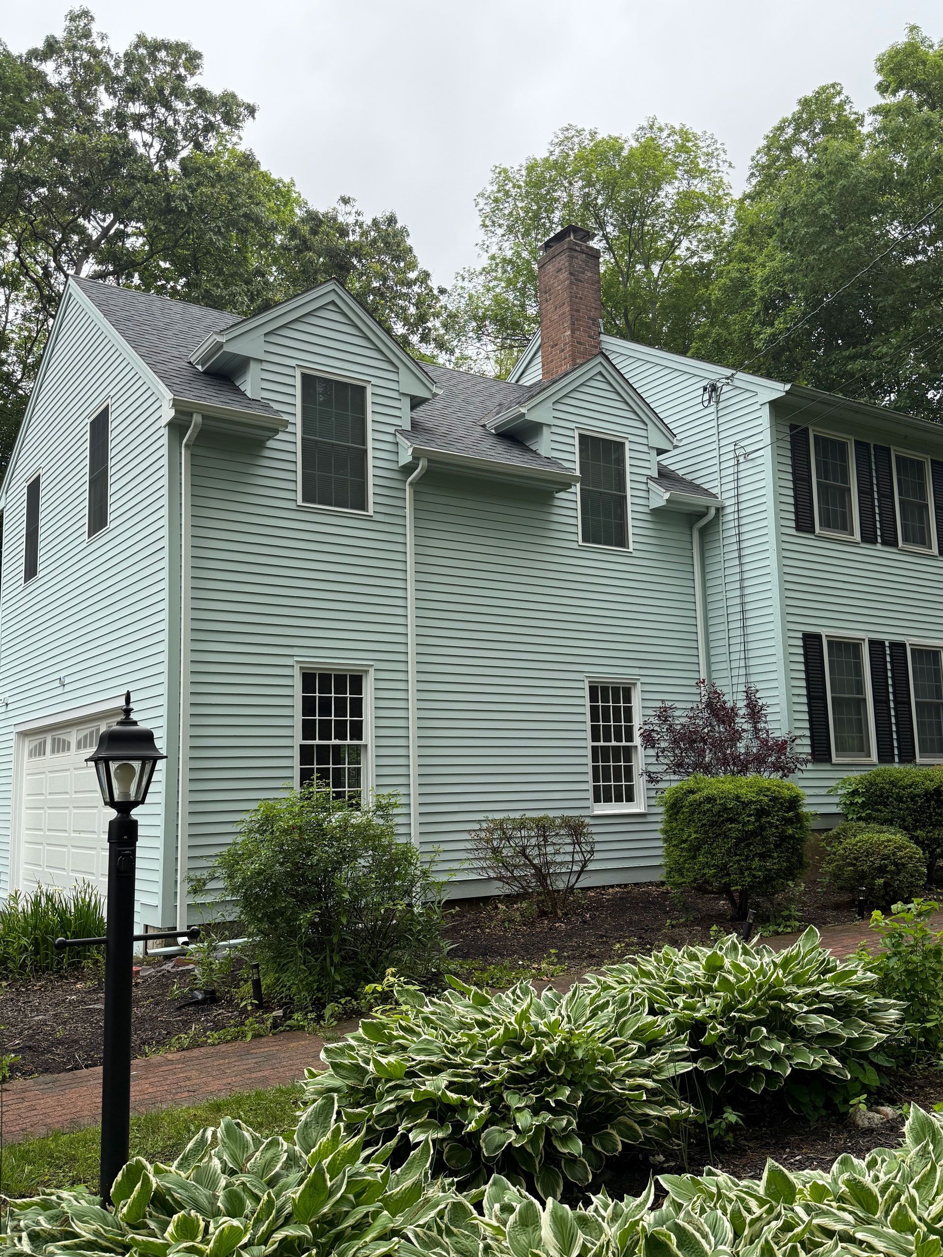 Mint-green house with a textured exterior, dormer windows, and a brick chimney behind shrubs and trees