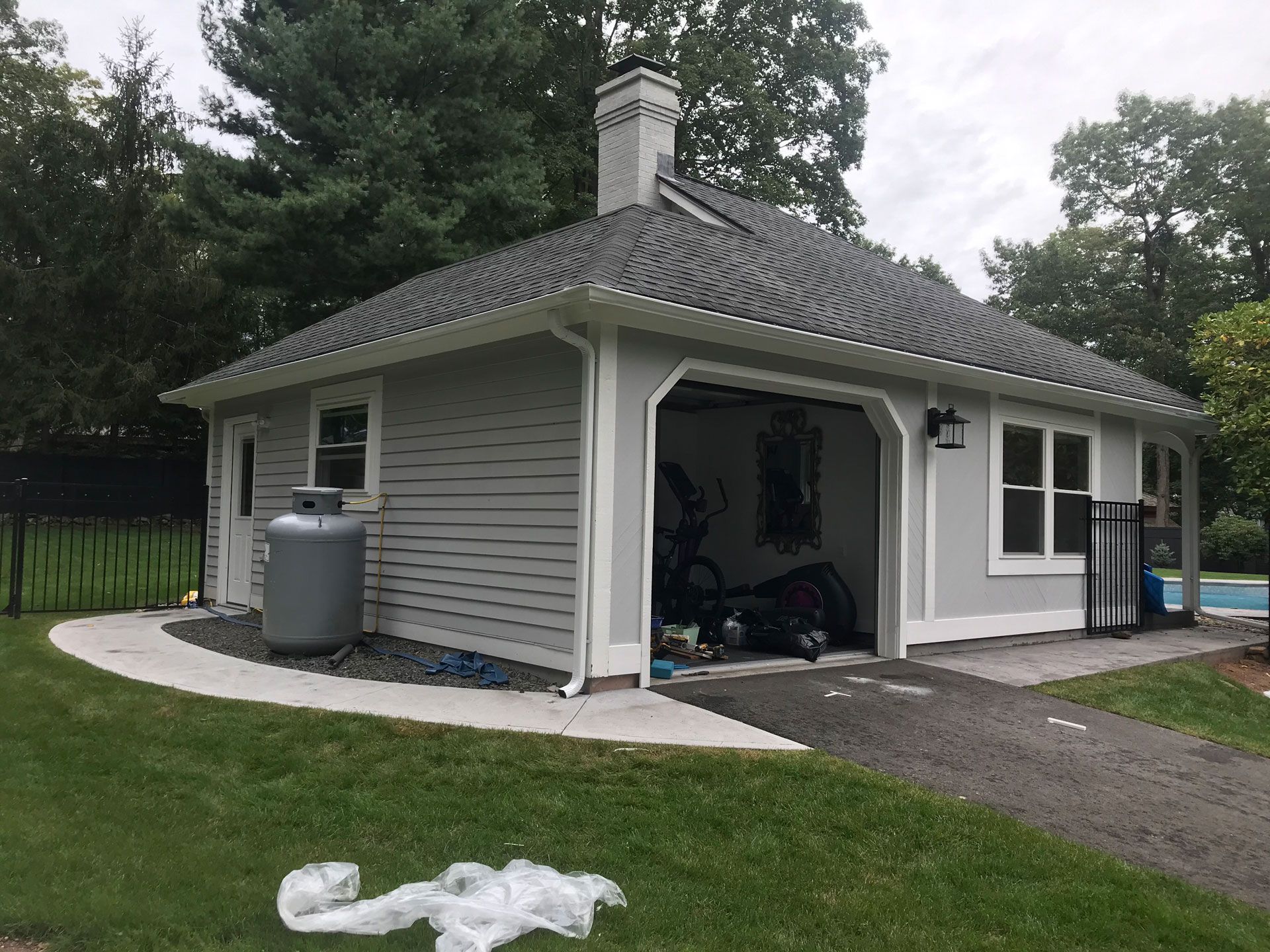 Small gray garage with open bay, white trim, driveway, and trees in a yard.