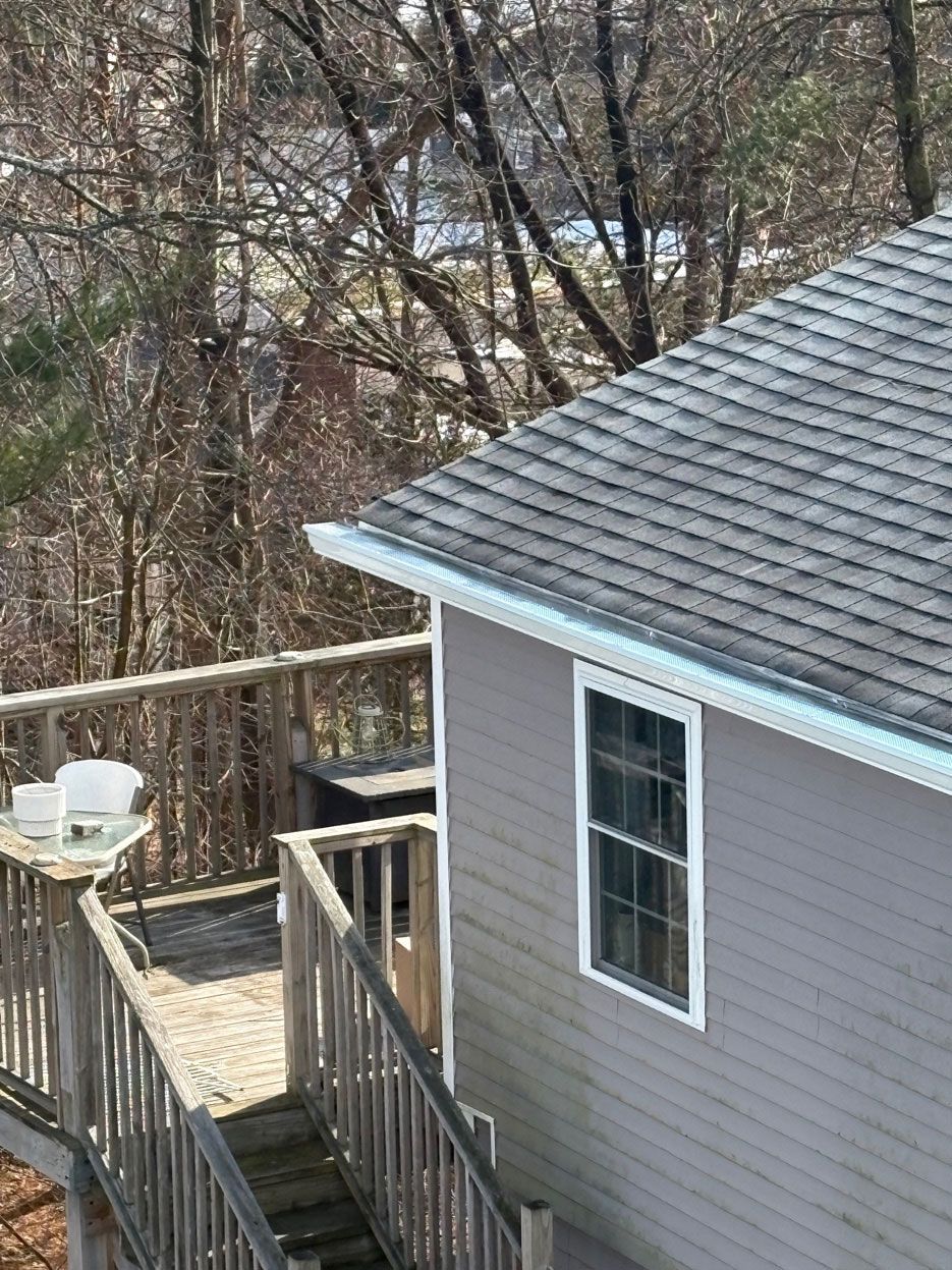 Wooden deck beside a gray house with a shingled roof, overlooking leafless trees and woods.