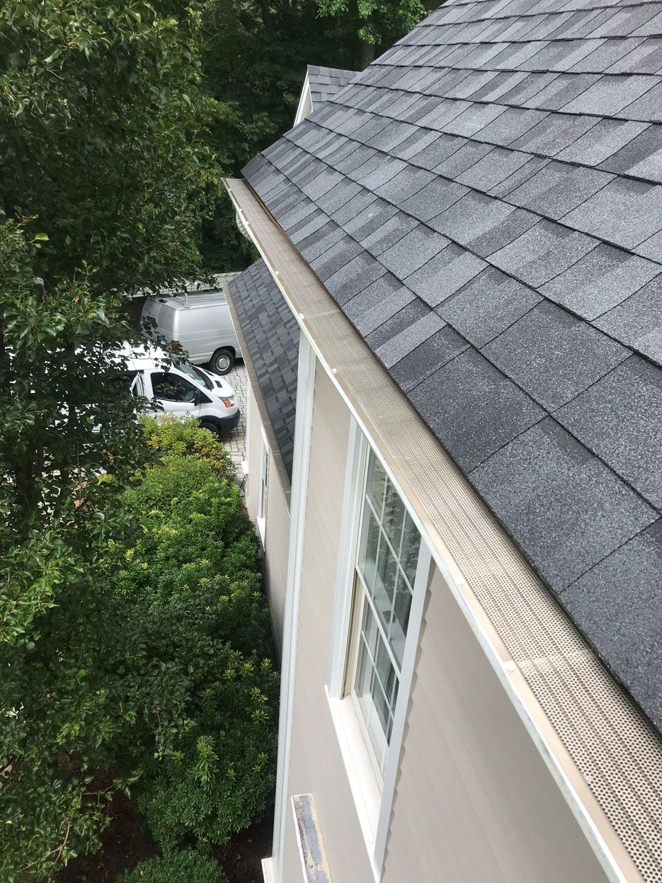 House roof and siding beside leafy trees, with a white vehicle partially visible below