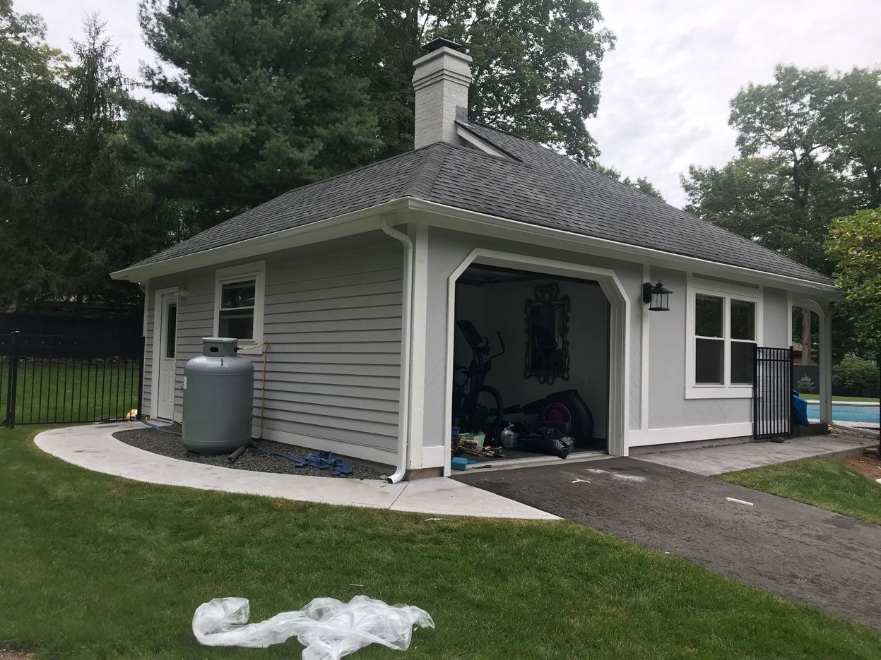 Small gray outbuilding with open garage door, white trim, and dark tiled roof beside a grassy yard.