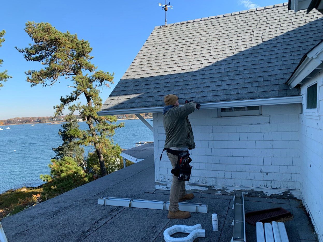 A person in work gear installs gutters on the edge of a white stone building roof overlooking a body of water.