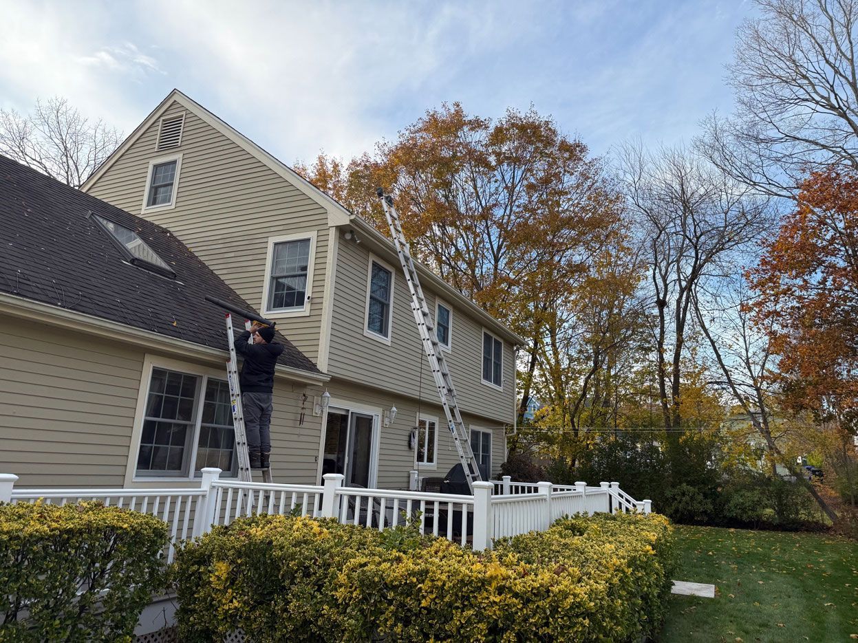 A person on a ladder repairs the exterior siding of a two-story beige house near trees in autumn.
