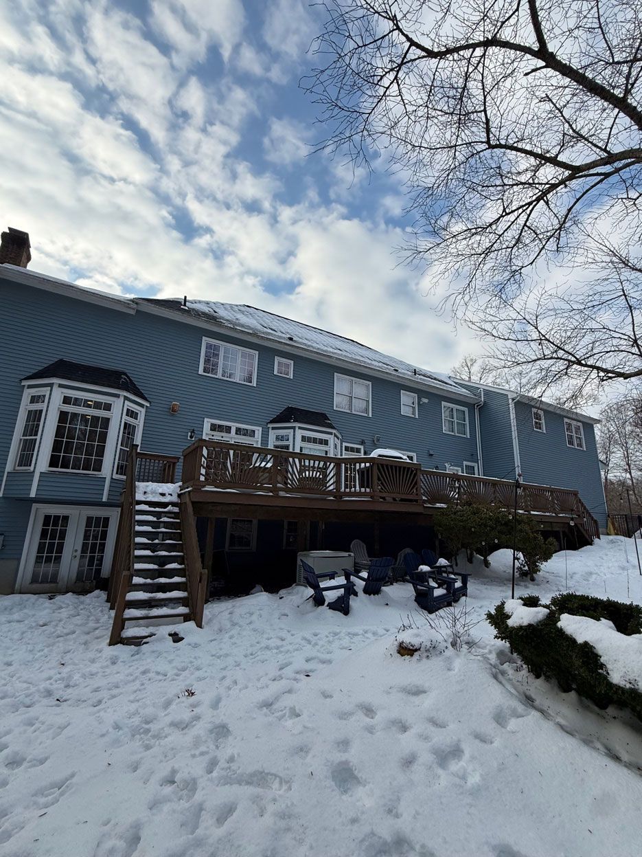 Snow-covered blue house with deck and steps, seen from the backyard under a cloudy winter sky