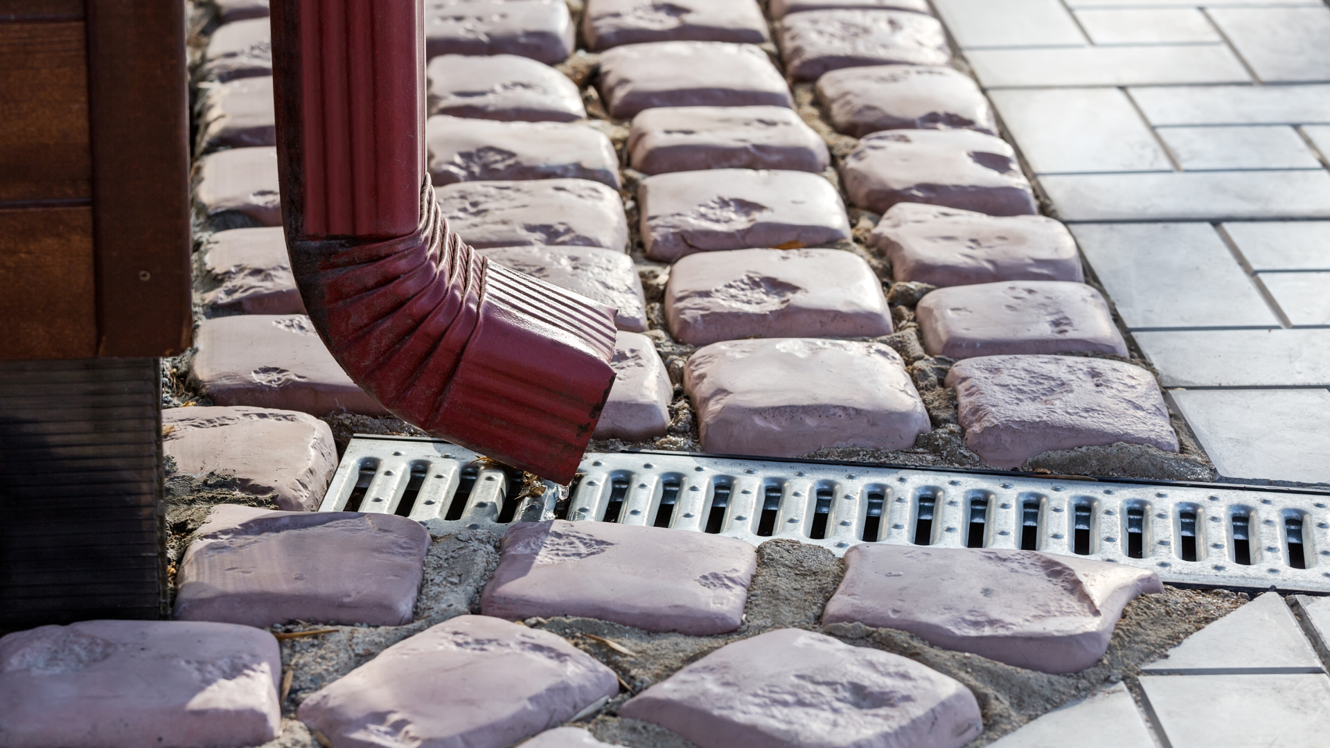 Rain gutter draining into a metal grate beside cobblestone pavers and tiled pavement