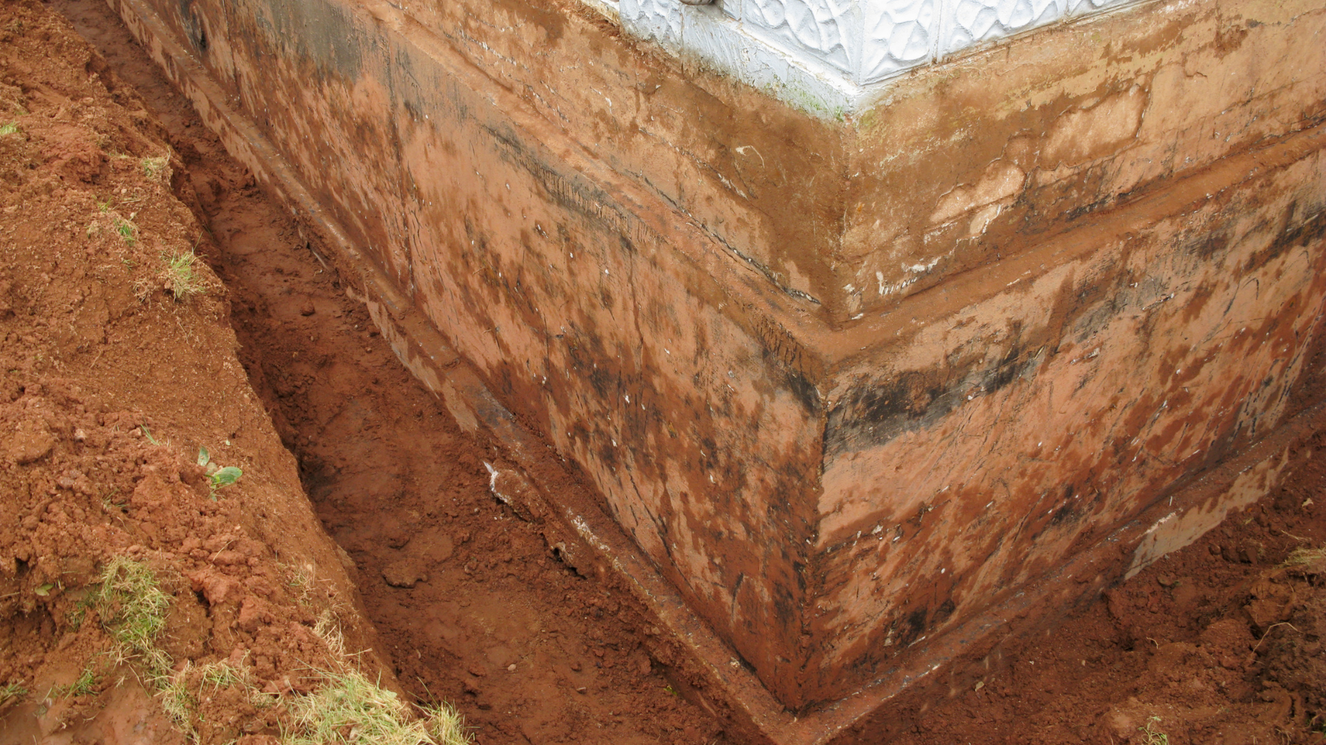 Corner of a white building with exposed brick foundation beside muddy soil.
