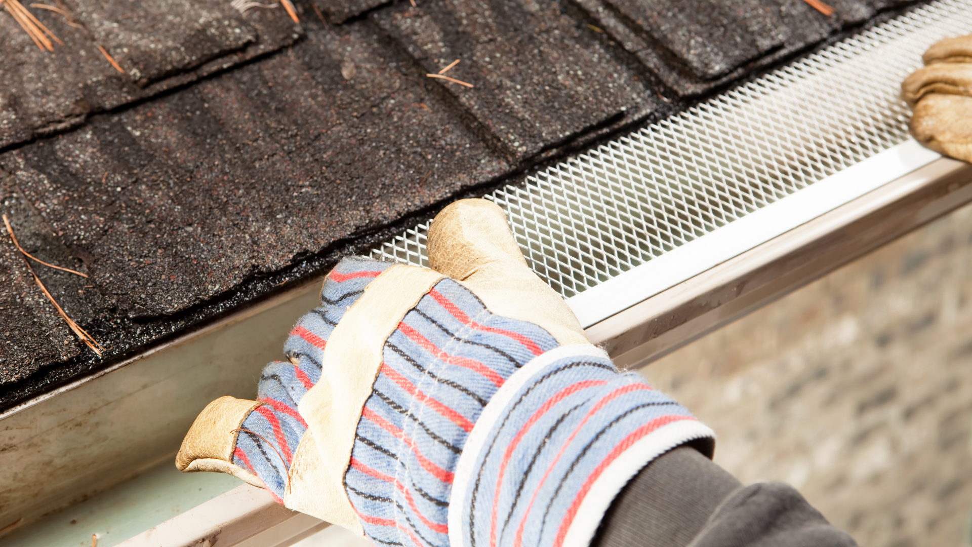 Gloved hands cleaning a rain gutter beside a shingled roof.