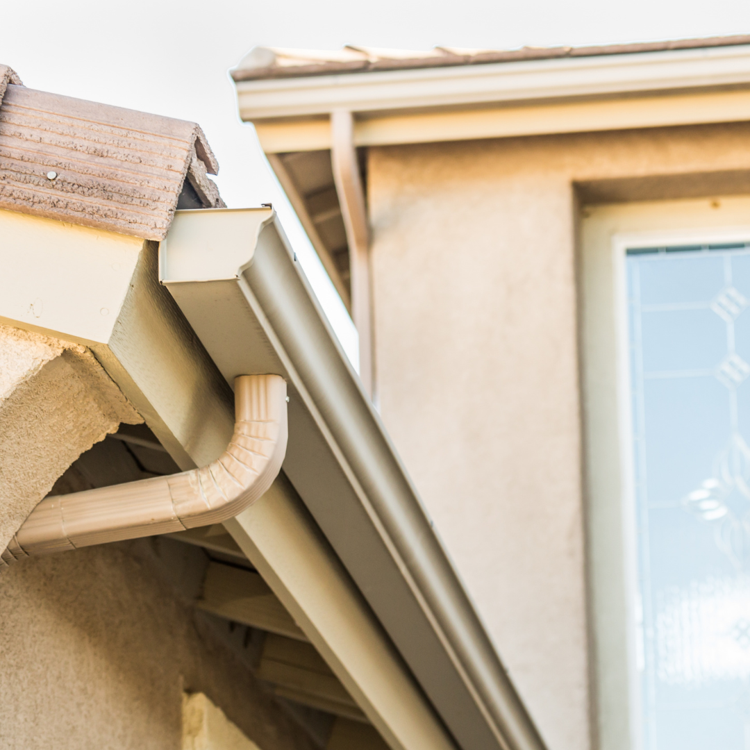 Close-up of house eaves with beige gutters and downspout beside a window