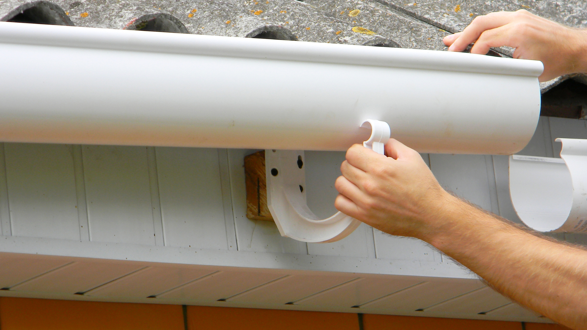 Hands installing a white gutter on a house roof edge