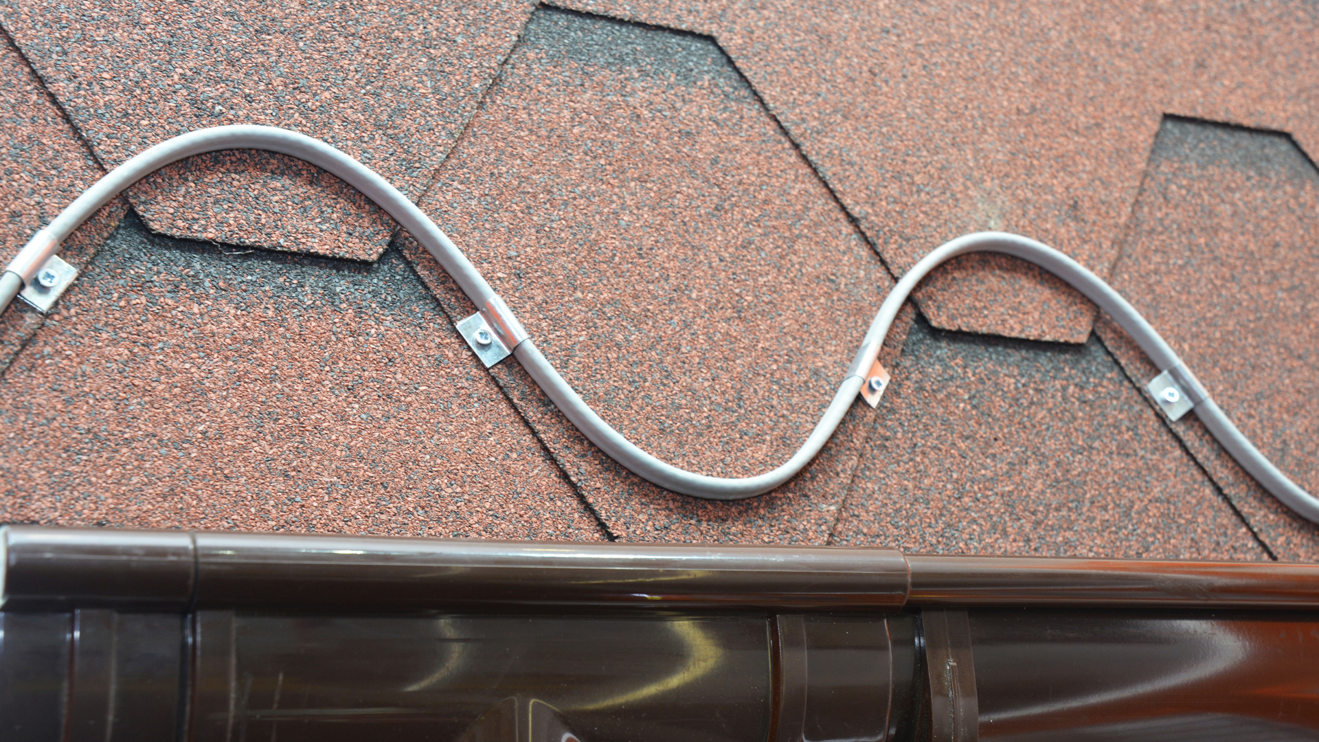 Coiled gray cable secured along a reddish shingle roof edge above brown guttering