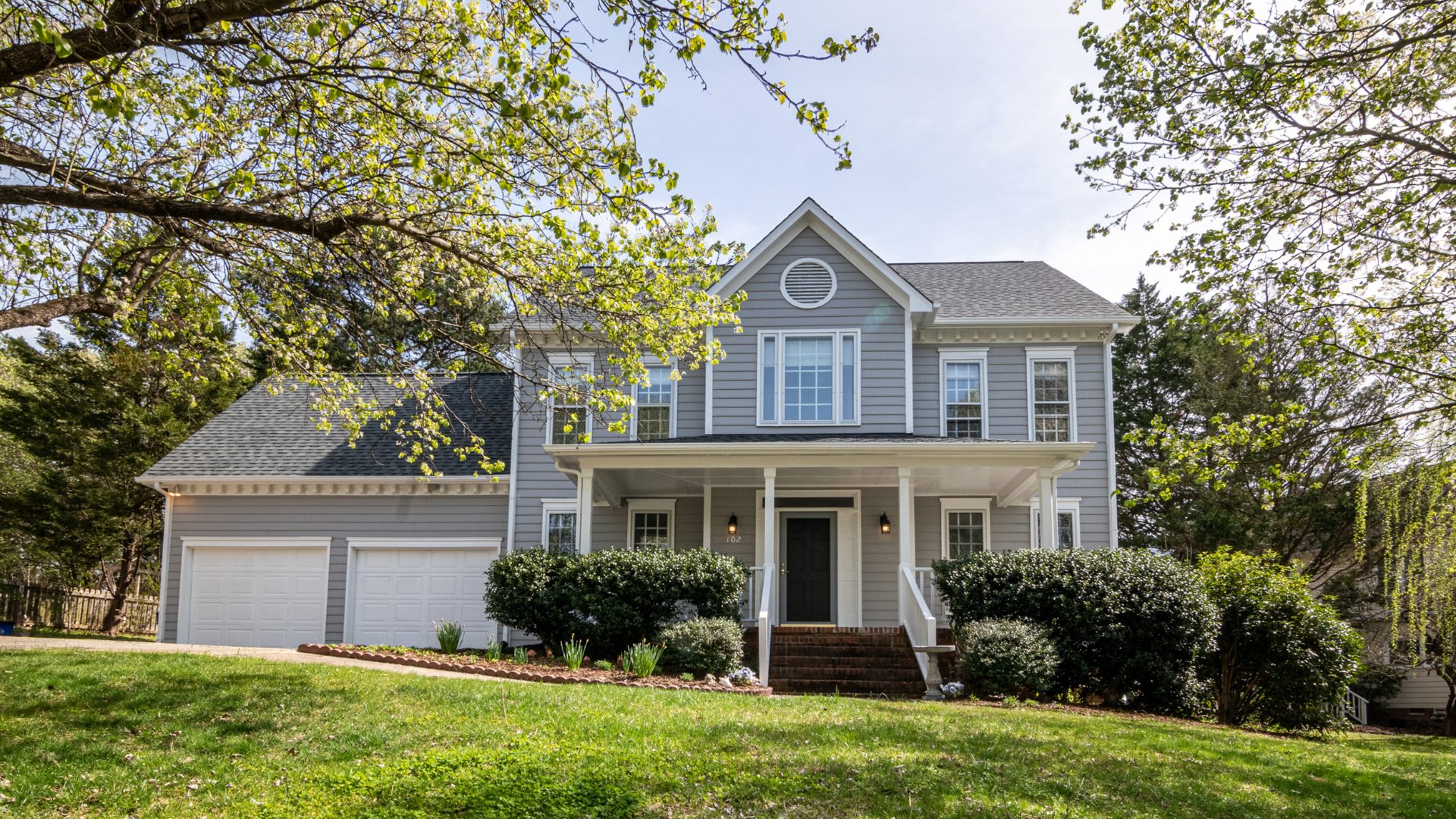 Suburban two-story gray house with white trim, front porch, garage, and trees in bloom