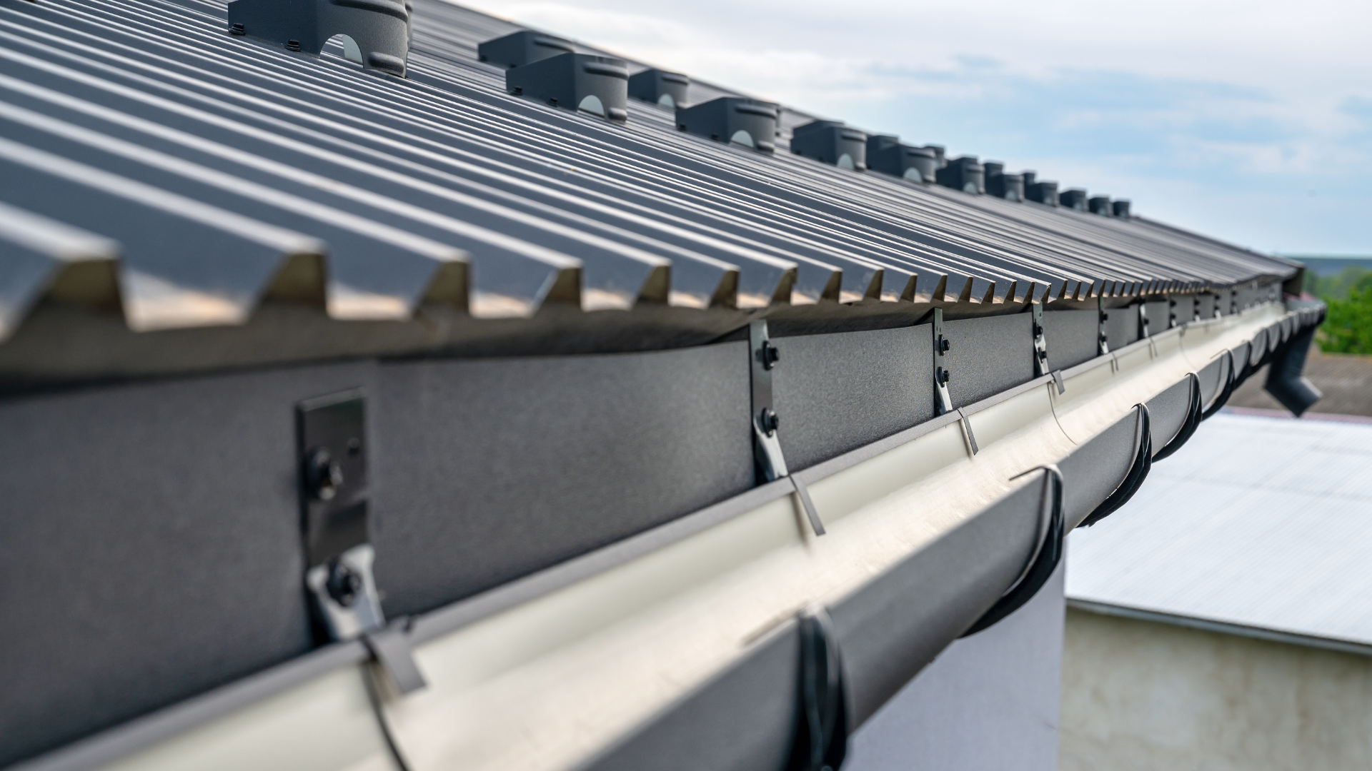 Person in a red jacket on a ladder cleaning a roof gutter against a blue sky