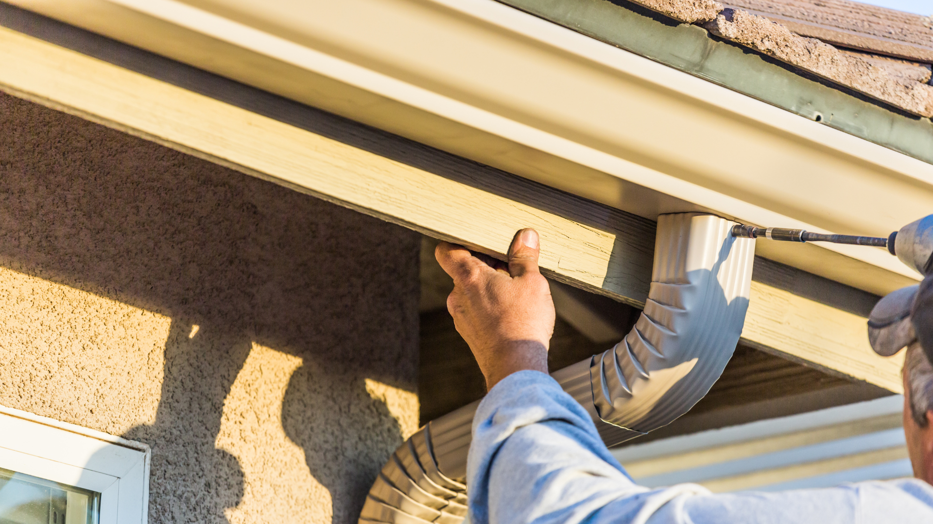 Worker installing or inspecting white rain gutters on a house roofline with a ladder nearby