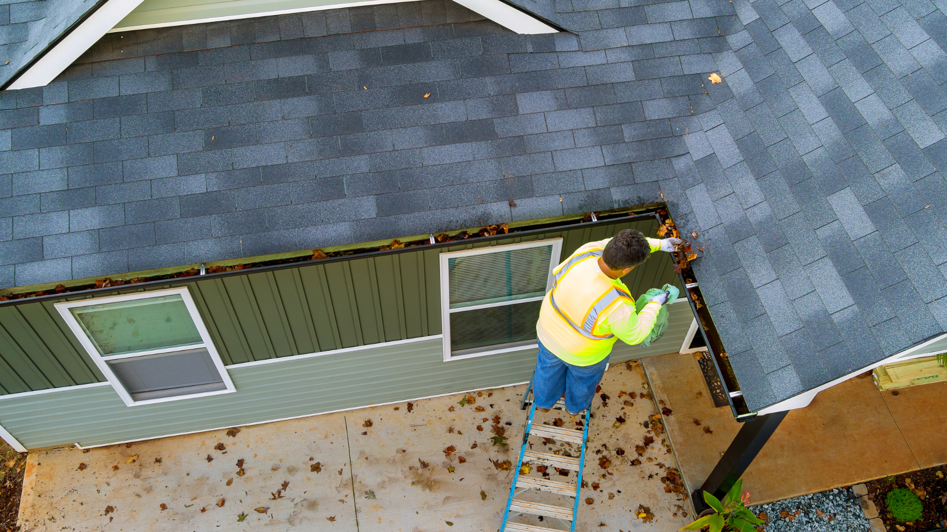 Worker on ladder repairing roof edge of a house with gray shingles.