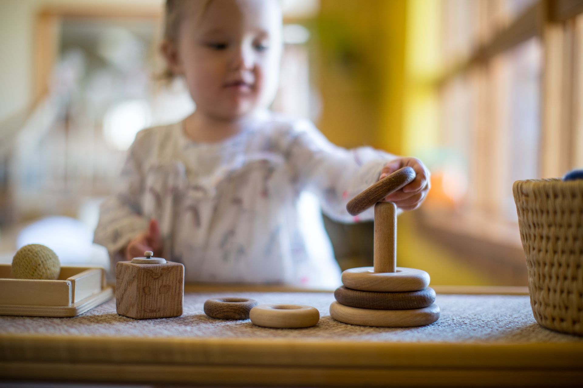 Toddler stacking wooden rings on a table with toys in a bright playroom