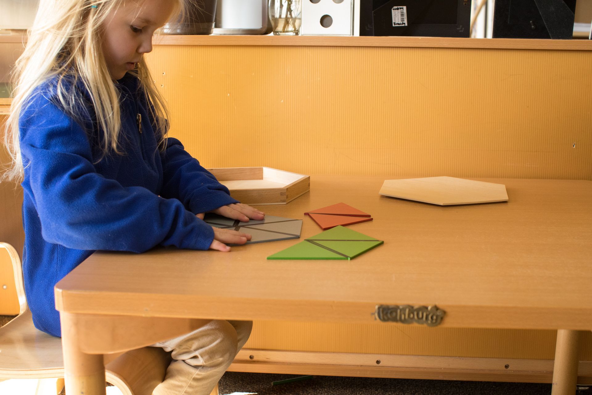 Child in blue sweater working with colorful geometric shapes at a wooden table in a classroom.