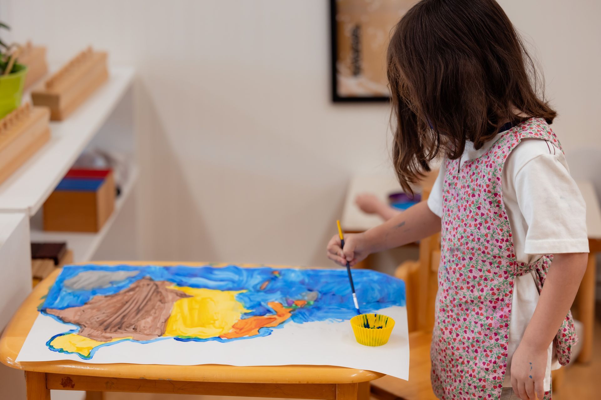 Child painting at a table with bright blue, yellow, and brown colors in a room with a shelf nearby