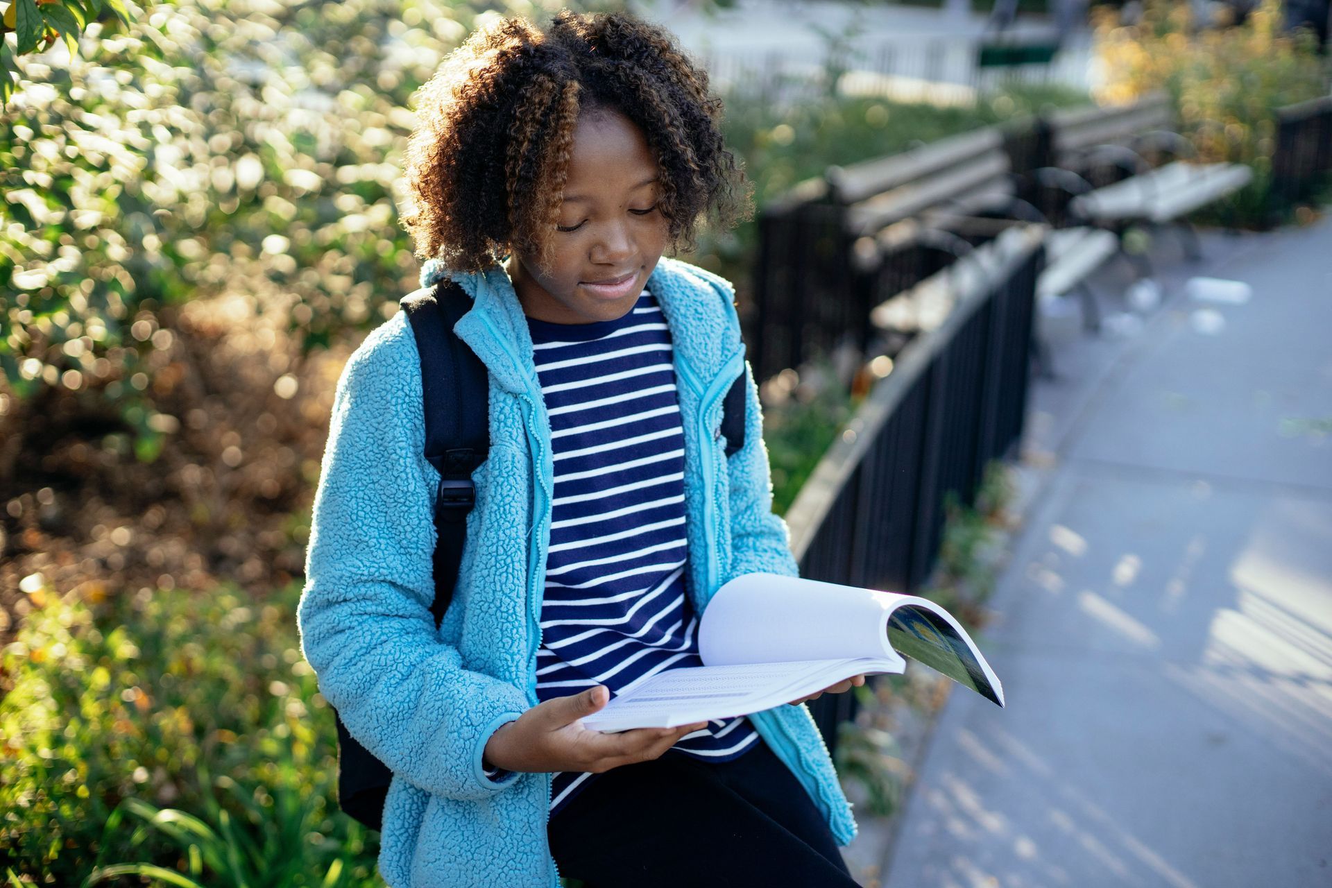 Person reading a book while walking along a sunlit path with a backpack and blue vest