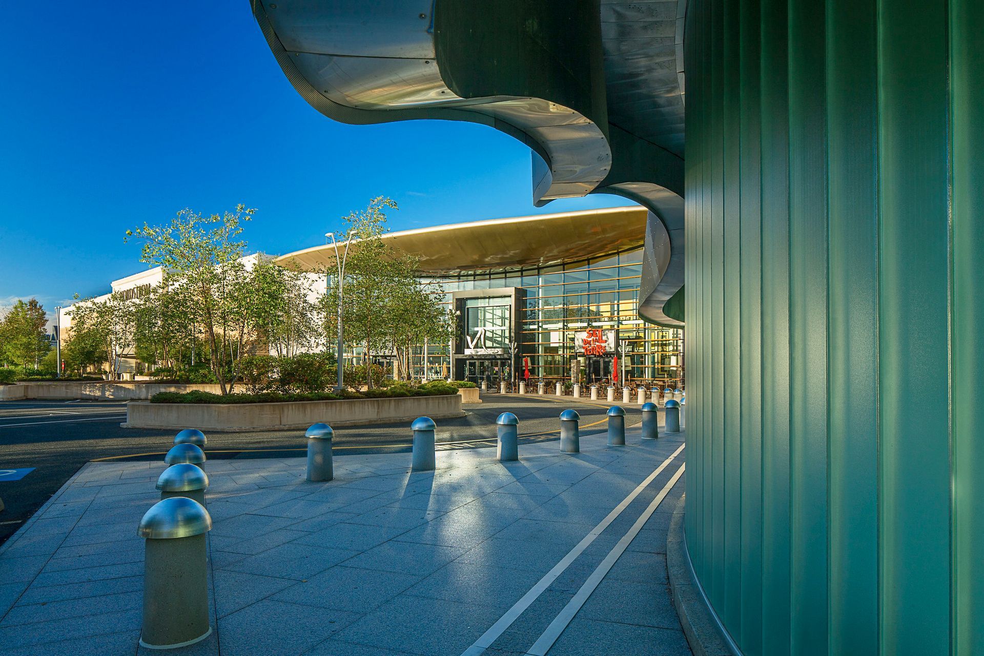 Modern building entrance with green columns, curved roof, and sunlit plaza under a blue sky