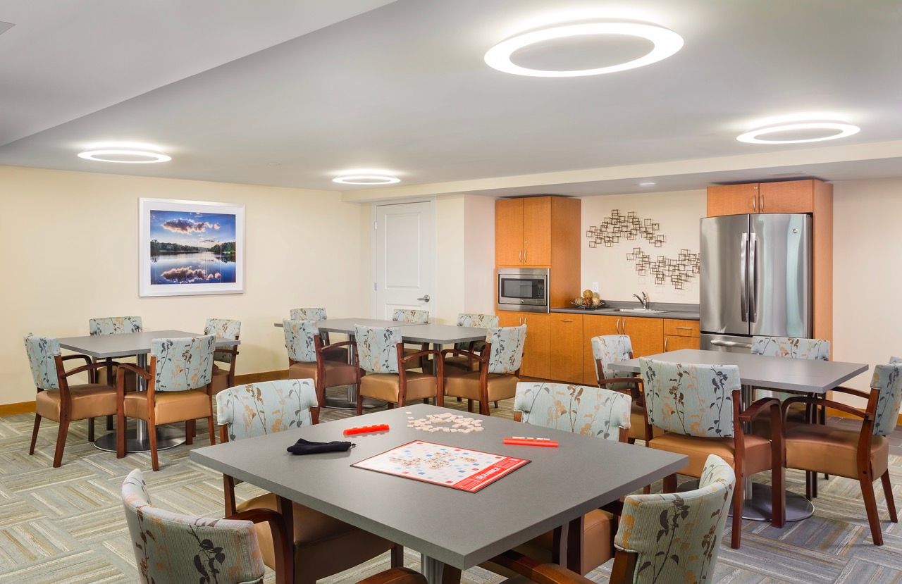 Bright community lounge with multiple tables, cushioned chairs, and a kitchenette along the back wall.
