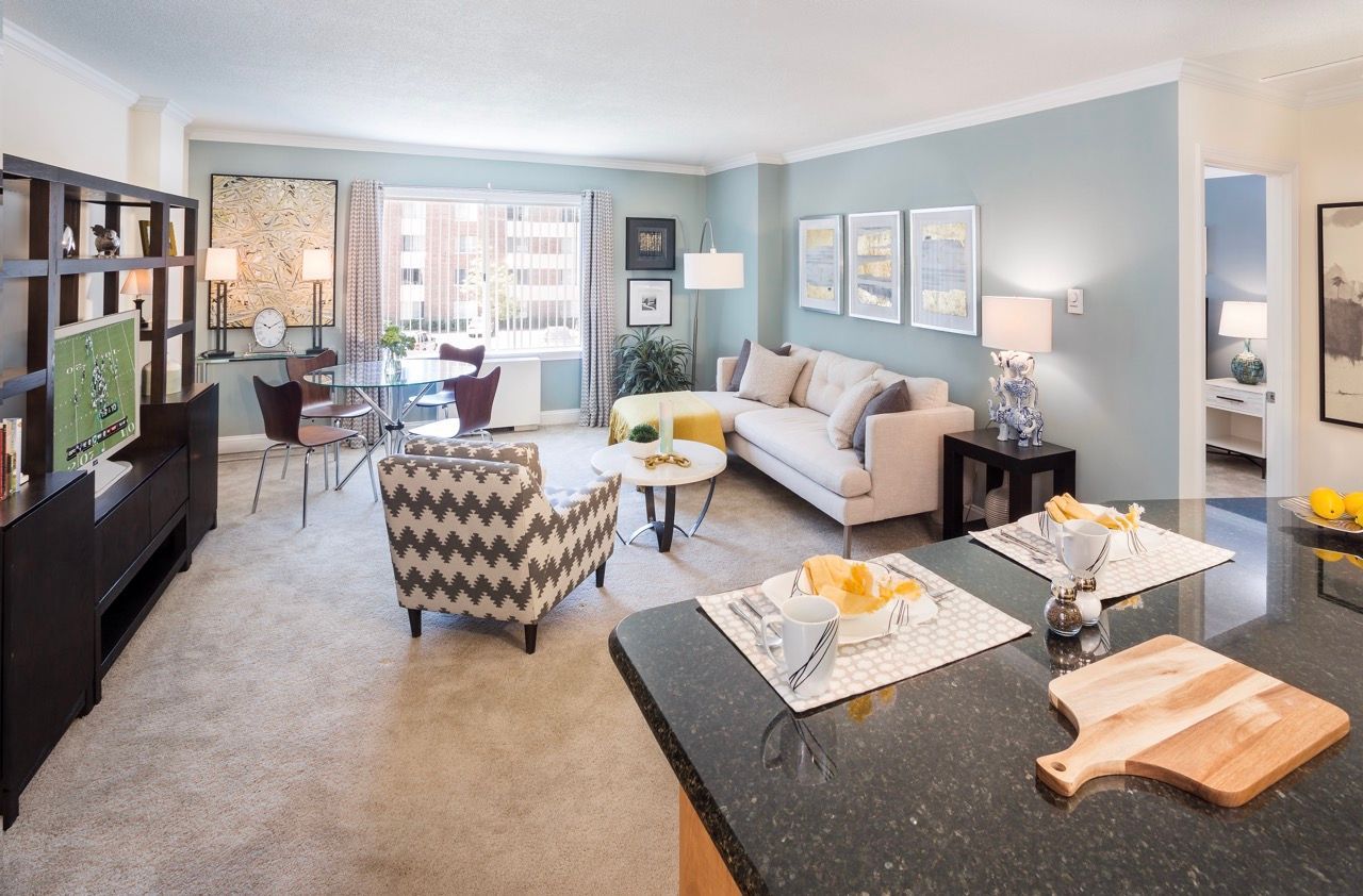Open-concept living area in a modern apartment with a beige sofa, patterned armchair, glass dining table, and blue accent wall.