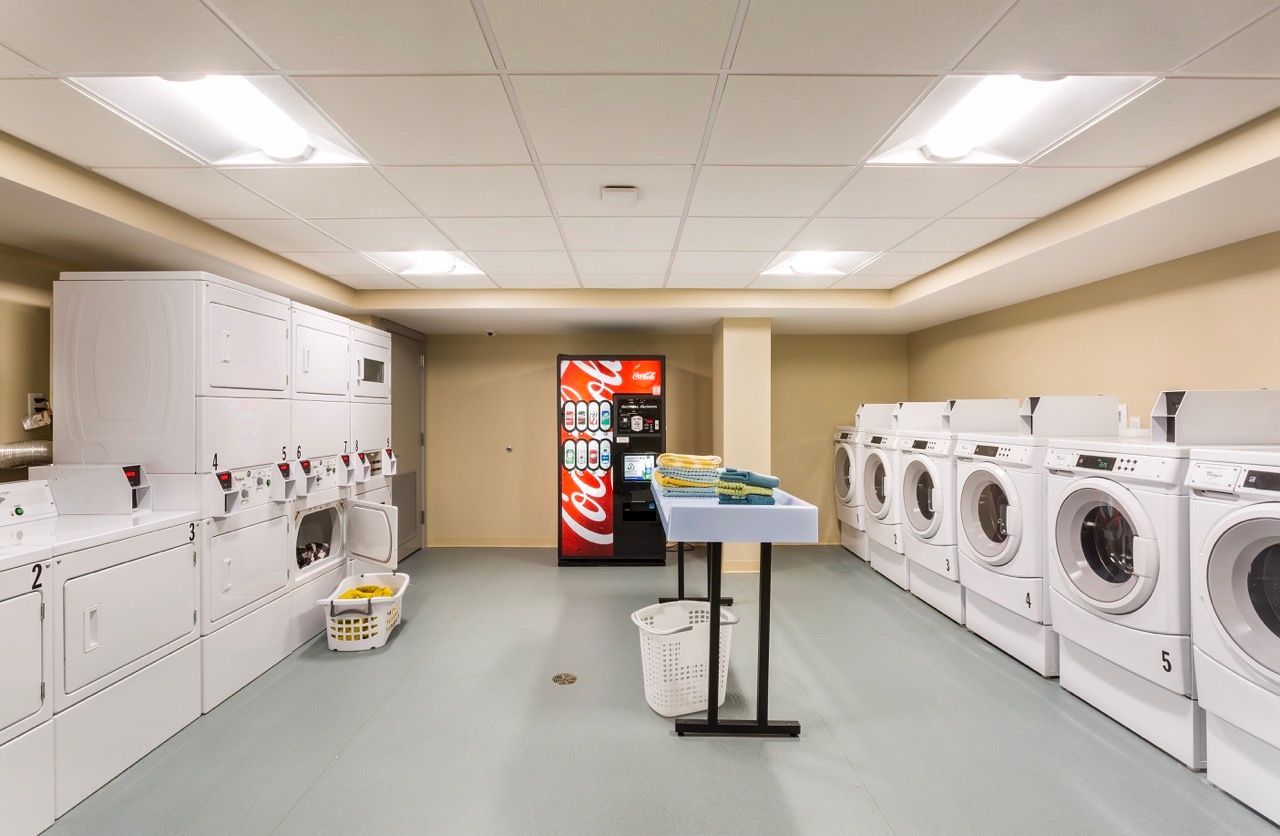 Communal laundry room with row of front-loading washers and dryers, a vending machine, and a folding table.