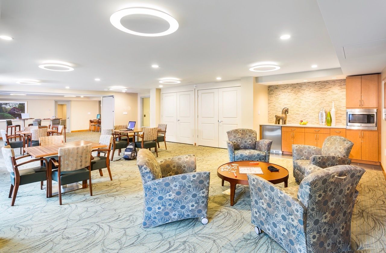 Indoor community lounge with beige walls, patterned chairs around tables, and a kitchenette.