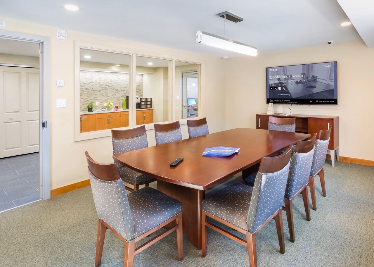 Conference room with a wooden table, eight upholstered chairs, and a wall-mounted screen.